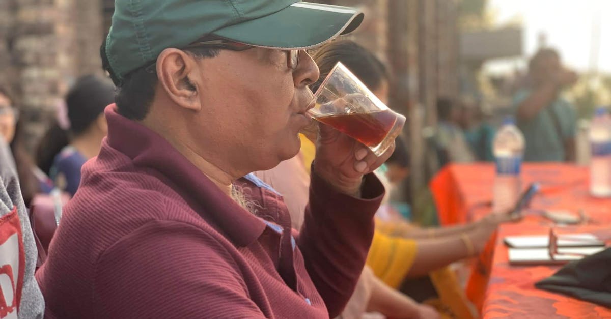 A man in a cap sipping a drink from a glass at an outdoor event during the day.