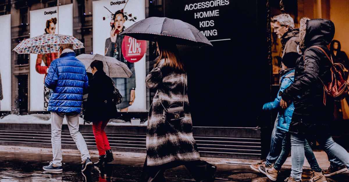 A bustling urban street with people walking in the rain, carrying umbrellas, and passing shop displays.