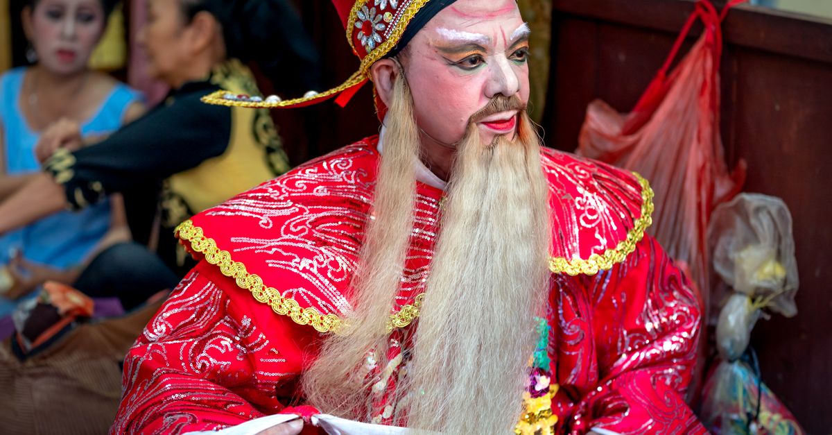 A Chinese opera performer in a red costume prepares backstage with detailed makeup.