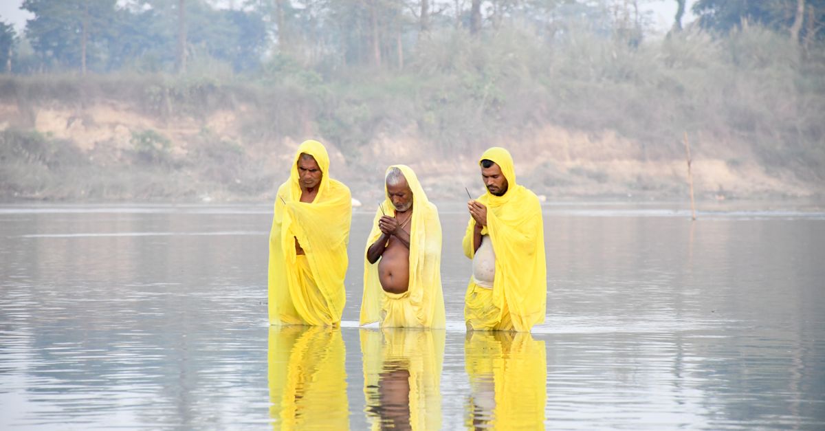 Three men in yellow garments perform a serene religious ritual standing in calm river waters.