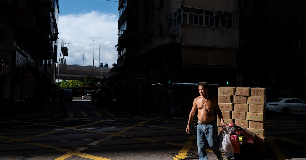 A man pulls a cart with boxes through a busy urban street intersection under bright daylight.