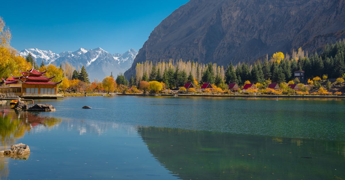 Peaceful autumn scene of a lake and mountains in Skardu, with vibrant foliage and a clear sky.