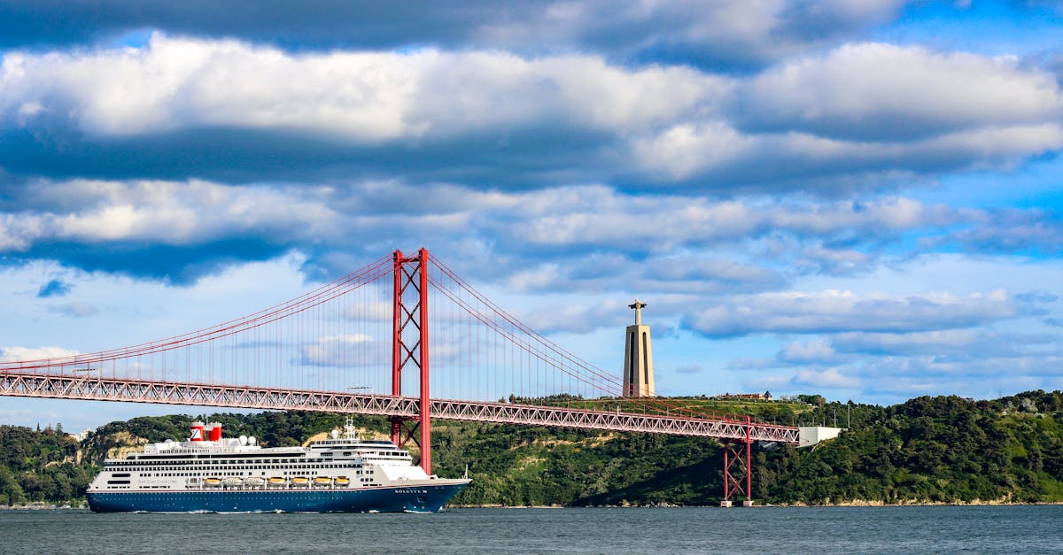 Scenic view of a cruise ship sailing under the iconic 25 de Abril Bridge in Lisbon with Christ the King statue in background.