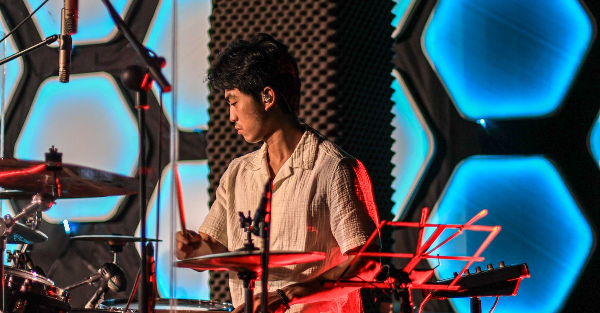 An Asian drummer playing a drum kit during a live concert with colorful lighting.