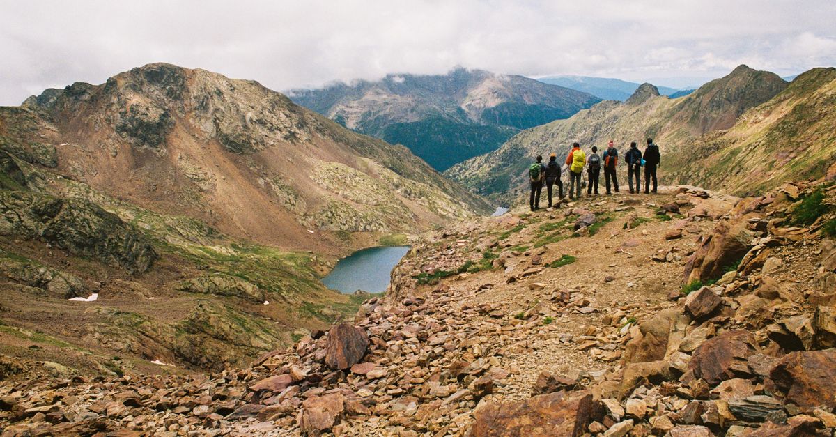 Group of hikers atop a mountain overlooking a scenic valley with a lake in the distance.