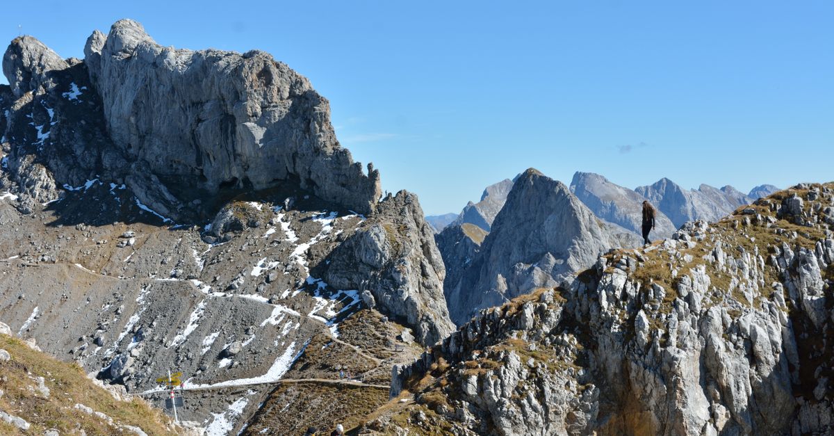 A lone hiker traverses a rugged mountain landscape against a clear blue sky.