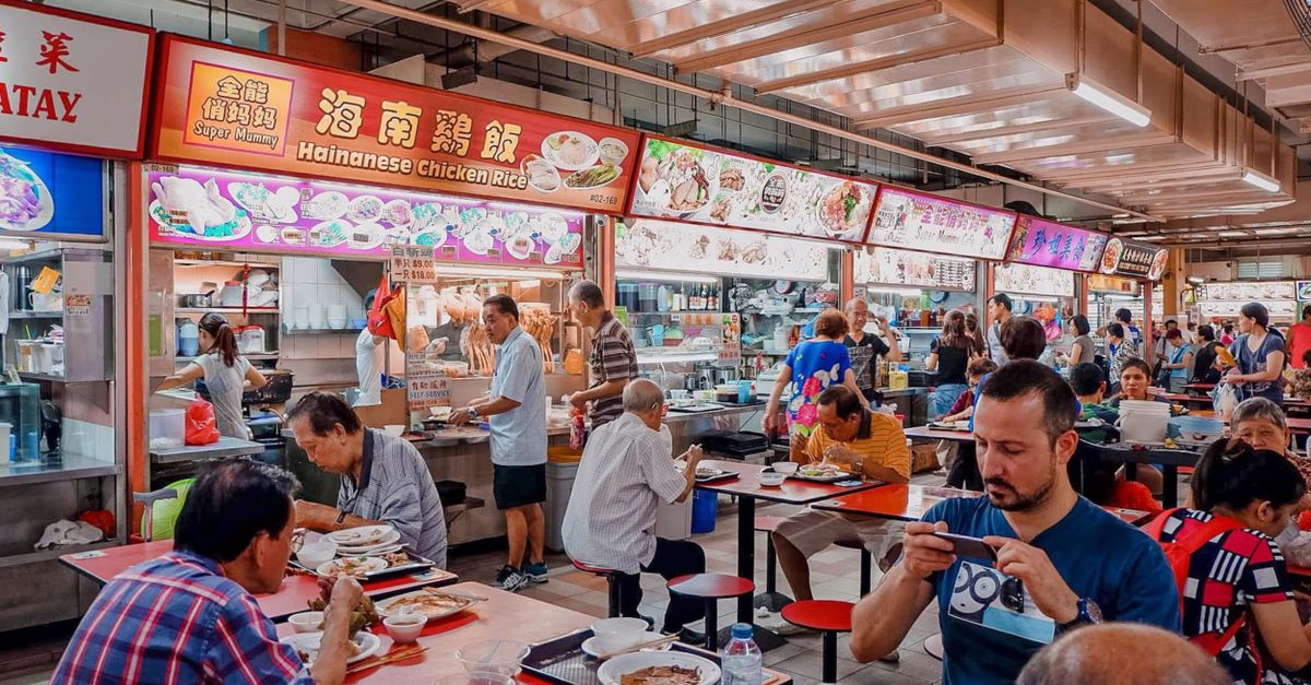 A bustling hawker center with diverse diners enjoying Hainanese chicken rice and various dishes.