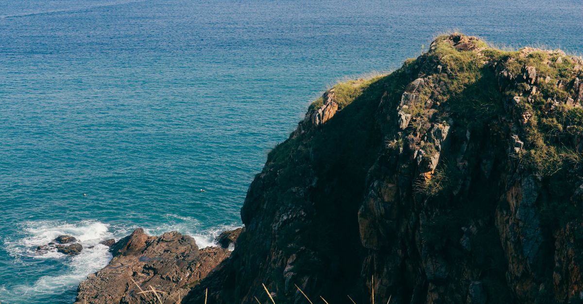 Majestic rocky cliff with lush grass overlooking a serene blue ocean under clear skies.