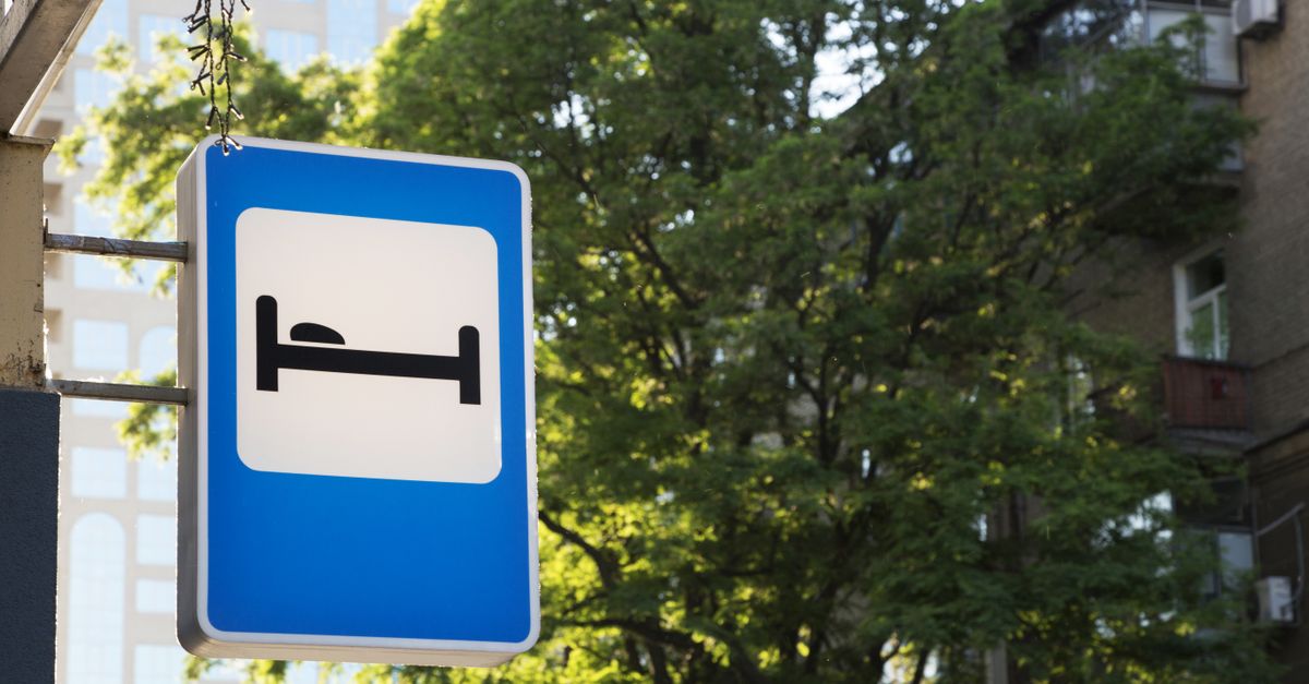 Hotel sign with bed symbol amidst urban greenery and buildings.