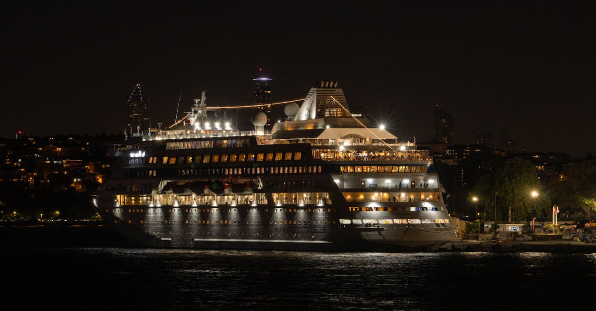 Illuminated cruise ship docked at night with city lights in the background, reflecting on water.