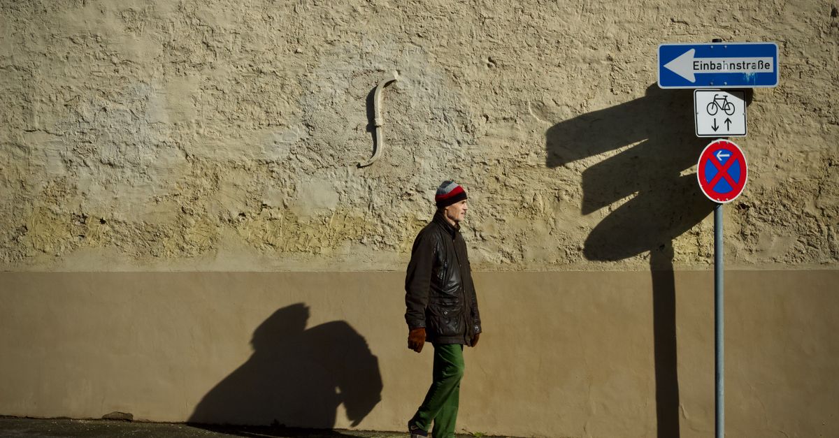 Man walks by wall under street signs casting shadows in urban setting.