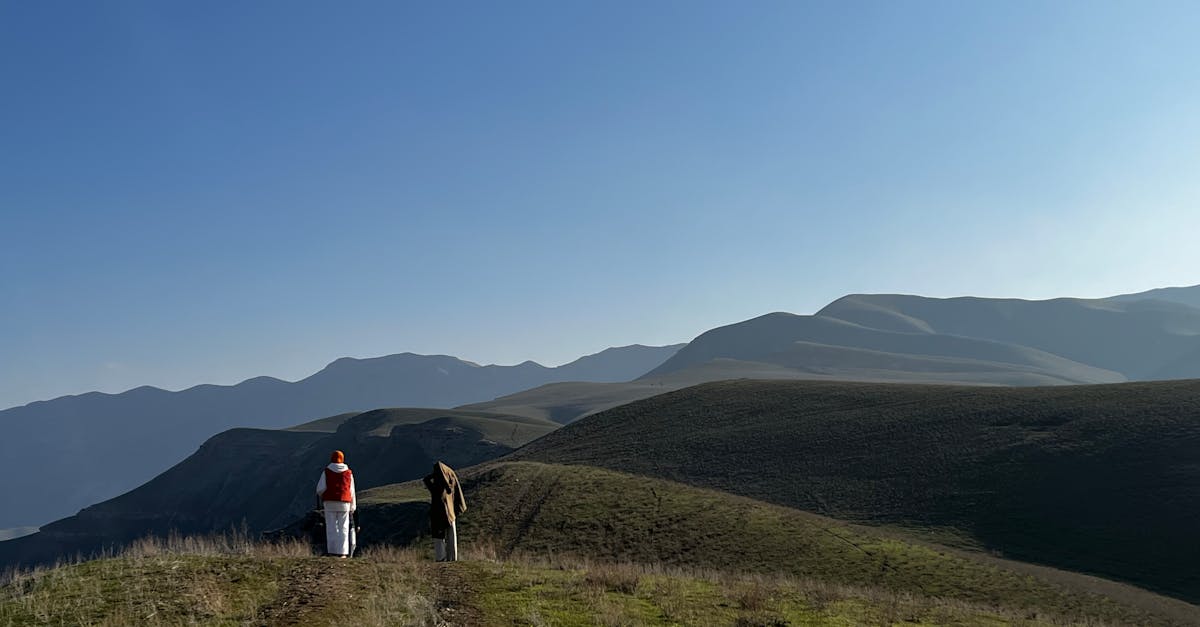 Two hikers explore a vast, rolling mountain landscape under a clear blue sky.