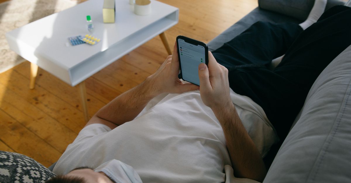 Man lying on a couch using smartphone with face mask and medication nearby, indoors.