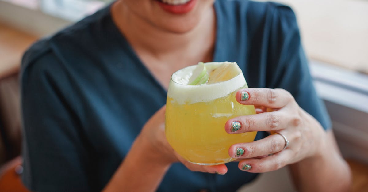 Cheerful unrecognizable female in casual wear sitting in restaurant near window with yellow cold drink with froth in hands on blurred background