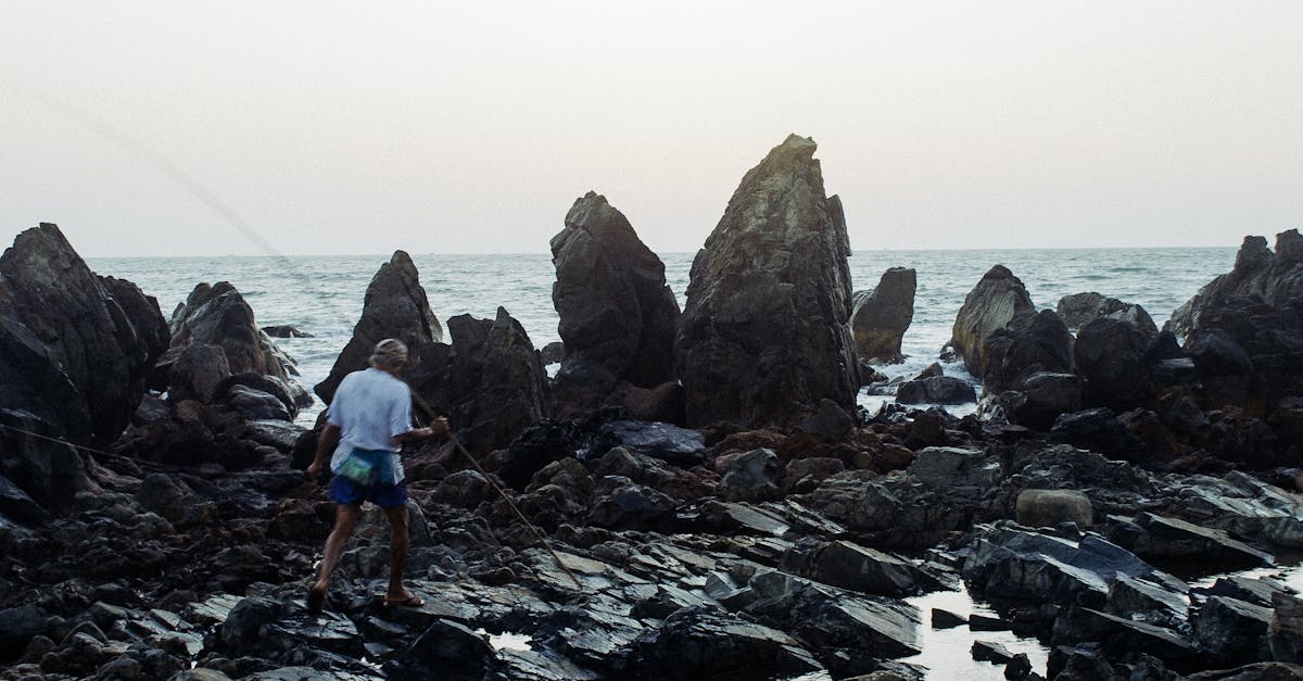 Back view full length local fisherman in casual summer wear carrying long bamboo stick and walking on rough stony seacoast on clear weather