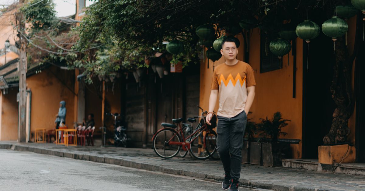 Full body of concentrated Asian man in casual outfit and glasses walking on city street near restaurant with Chinese lanterns and looking away at daytime