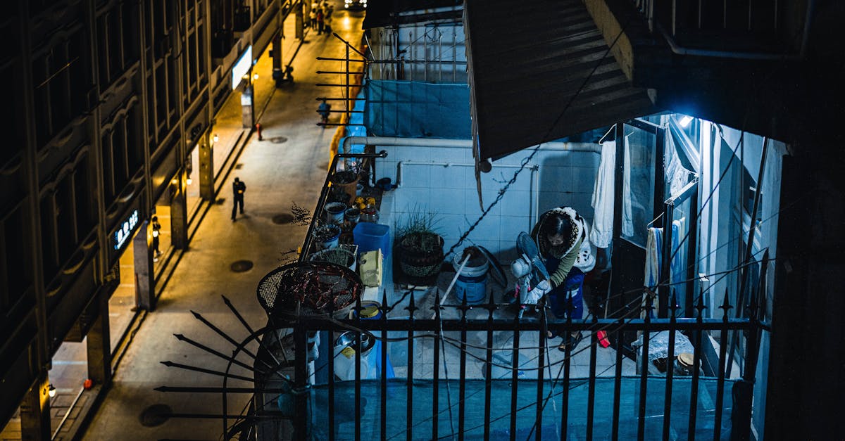From above of distant unrecognizable female putting fan on balcony with metal fence of residential buildings located on street at night time
