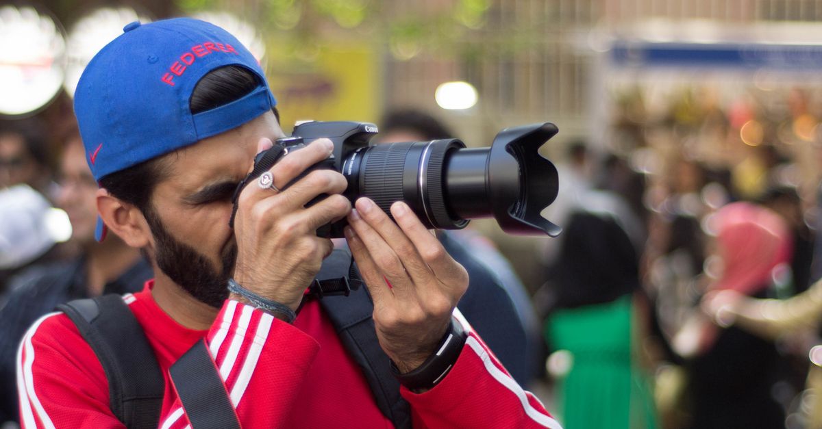 A photographer in a blue cap takes pictures at a busy outdoor event.