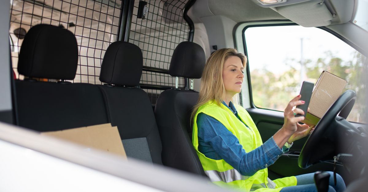 Female courier in safety vest holding parcel in van while using smartphone for delivery instructions.