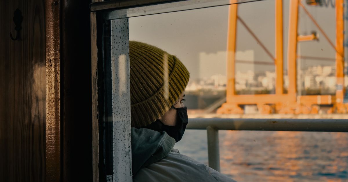 A person wearing a yellow beanie and face mask gazes over the sea from a ferry, deep in thought.