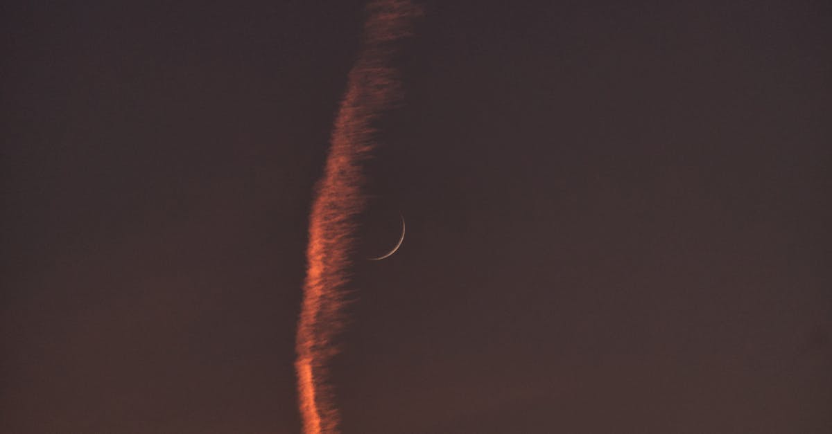 A serene view of a crescent moon beside a glowing contrail in the evening sky.